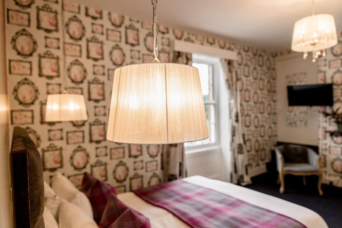 A bedroom with patterned wallpaper, a bed with a plaid blanket, and two hanging lamps.