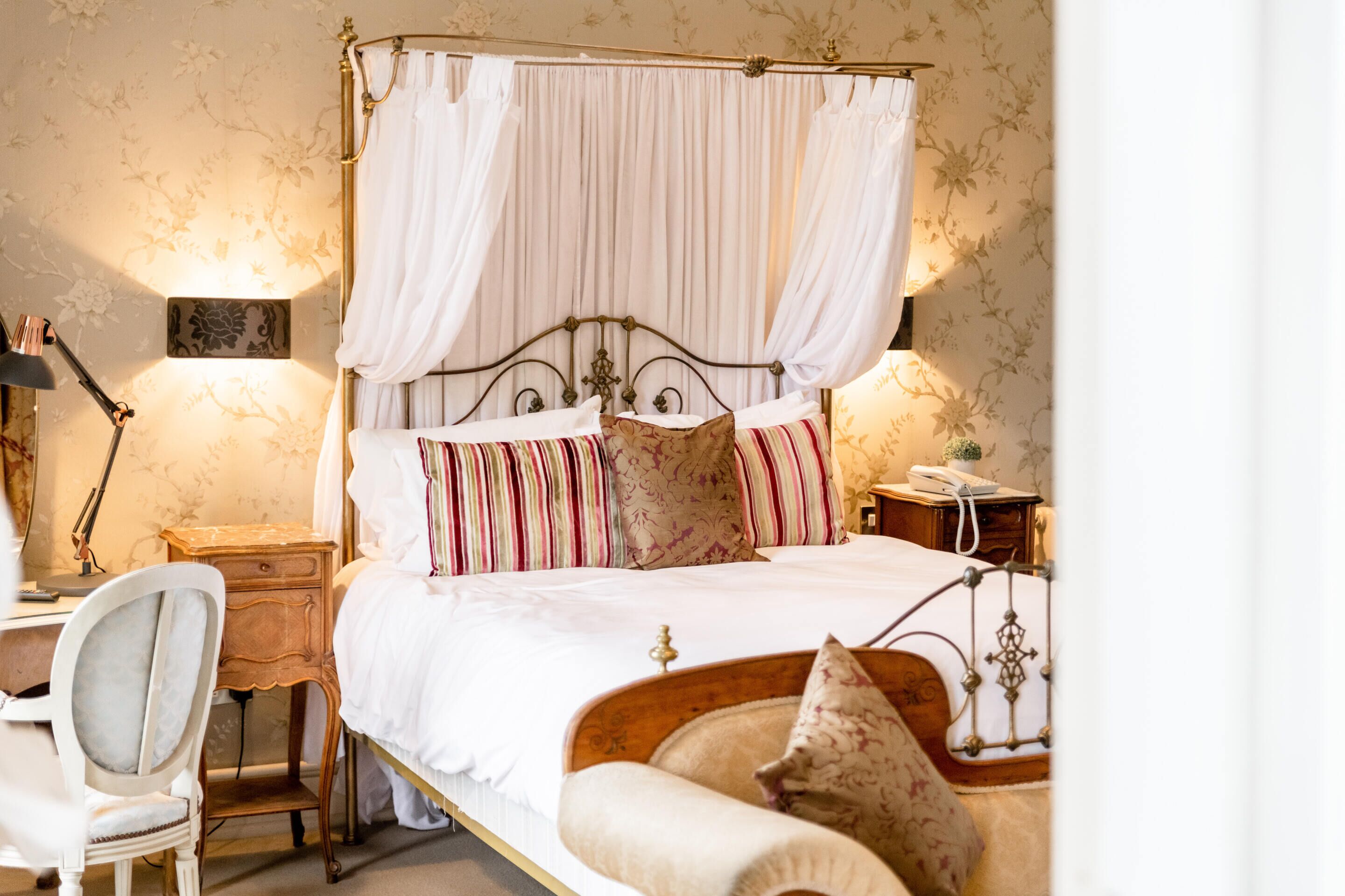 A four-poster bed with white drapes, a wooden nightstand with a lamp, a desk with a chair, and a telephone.