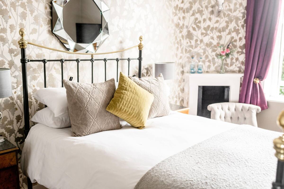 A neatly made bed with a headboard, a mirror above it, and a fireplace in the background.