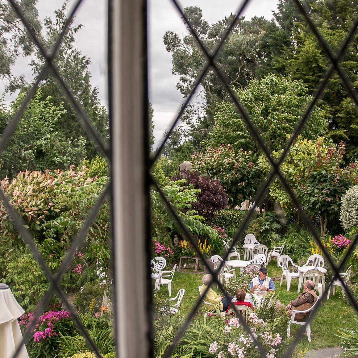 A garden with white plastic chairs and tables, surrounded by lush greenery and flowering plants.