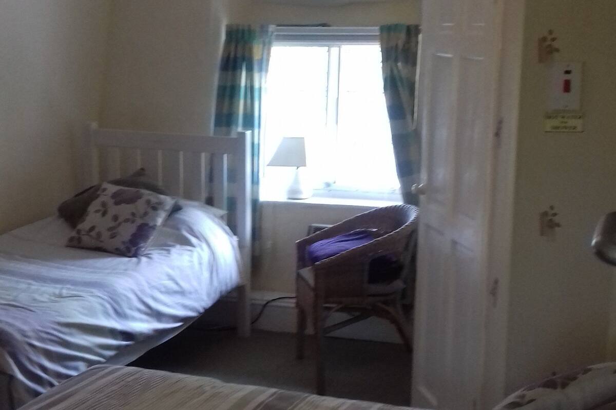 A small, cozy room with two single beds, a desk, and a chair. There is a window with curtains, a lamp on the desk, and a radiator.