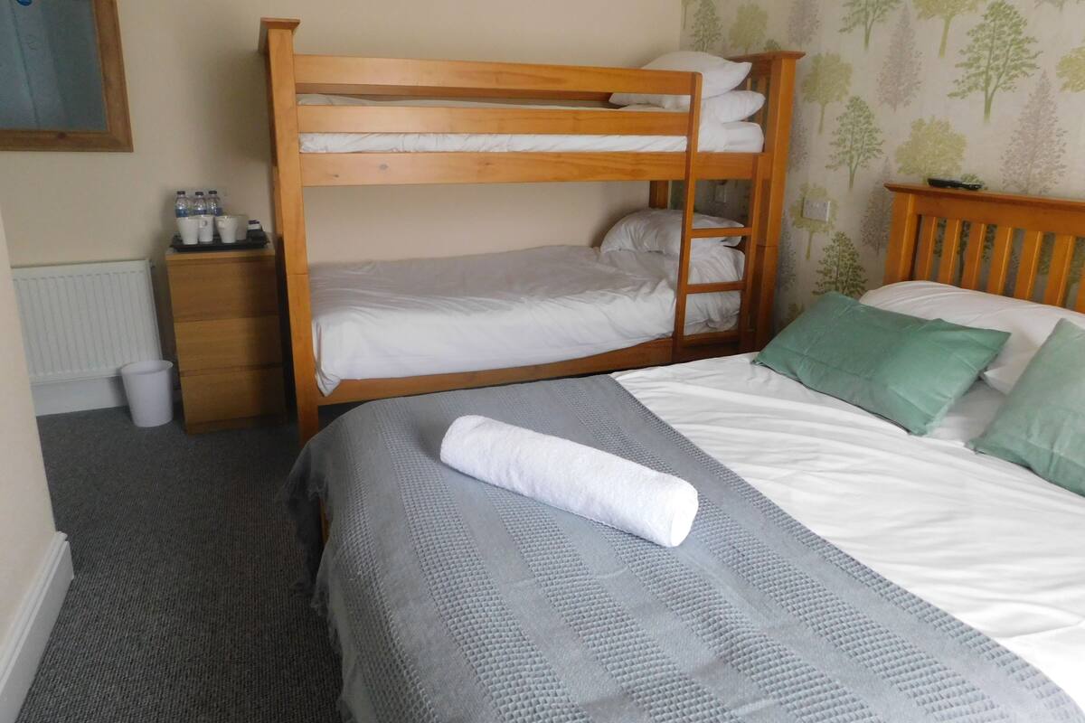 Een slaapkamer met stapelbedden, een houten hoofdbord, een klein houten nachtkastje met flessen, een grijs bed met een geruisige deken en een groen kussen.