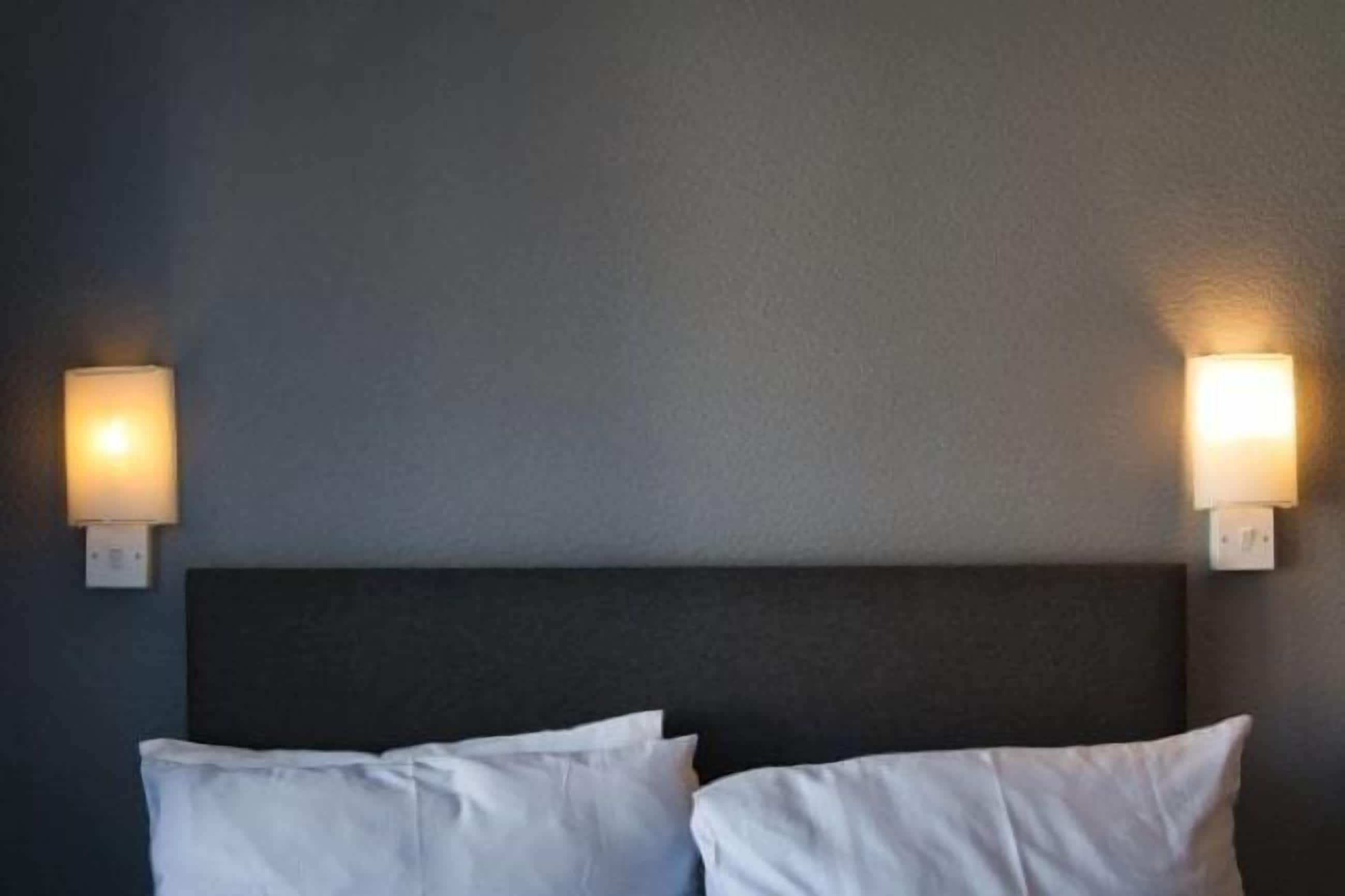 A hotel room with a dark headboard, two white pillows, and wall-mounted lights on either side.
