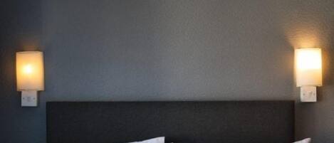 A hotel room with a dark headboard, two white pillows, and wall-mounted lights on either side.