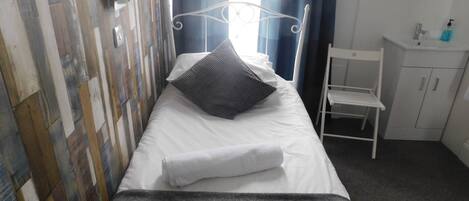 A single bed with a white mattress and a grey blanket, a grey pillow, and a folded white towel. There is a white metal headboard, a small white chair, and a white cabinet with a mirror above it. The wall has a wooden panel design.