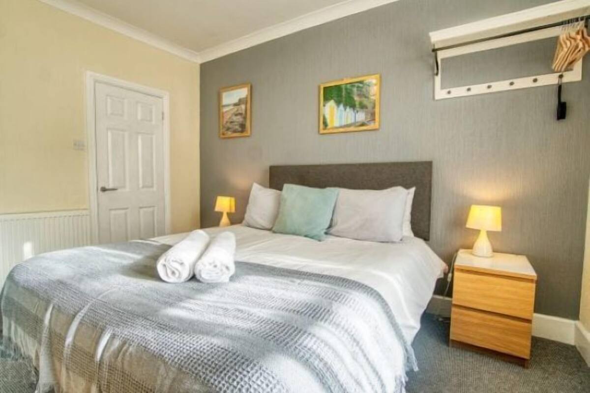 Een keurig opgemaakt bed met een grijs-witte deken, twee kussens en opgerolde handdoeken. Er staat een nachtkastje met een lamp en een klein lade. De kamer heeft een deur, een wandplank met kleding en twee ingelijste foto’s.