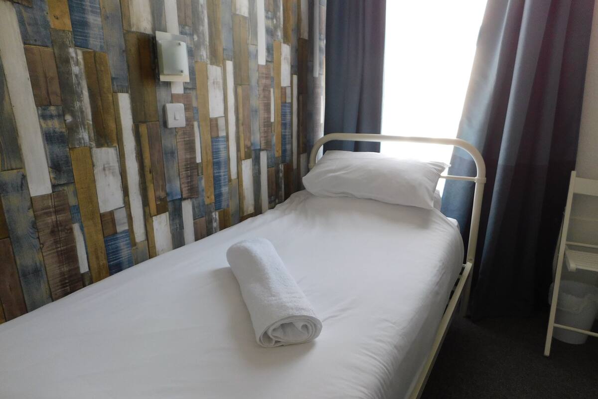 Een enkel bed met een opgerolde witte handdoek, een houten wand met een mix van planken, een witte wandlamp en een raam met gordijnen.