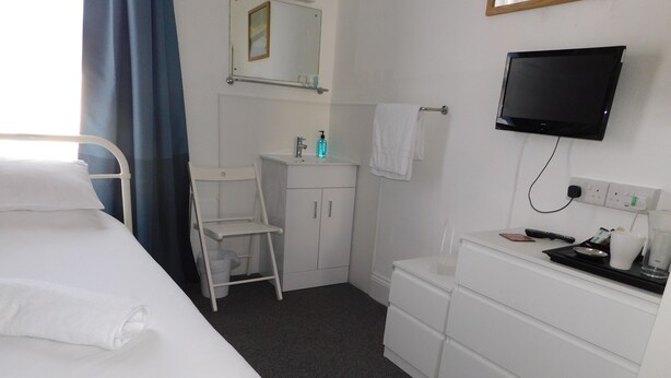 A hotel room with a bed, a white dresser, a TV mounted on the wall, a small bathroom with a sink and mirror, and a window with blue curtains. - St Weonards Hotel (Paignton)