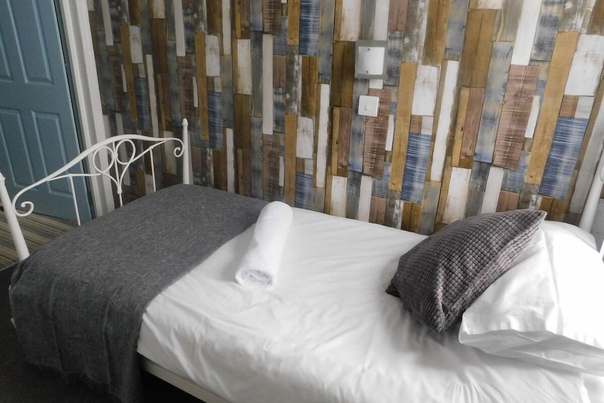 A single bed with a white mattress, a gray blanket, and a rolled white towel. The bed is against a wall with a wooden plank pattern.