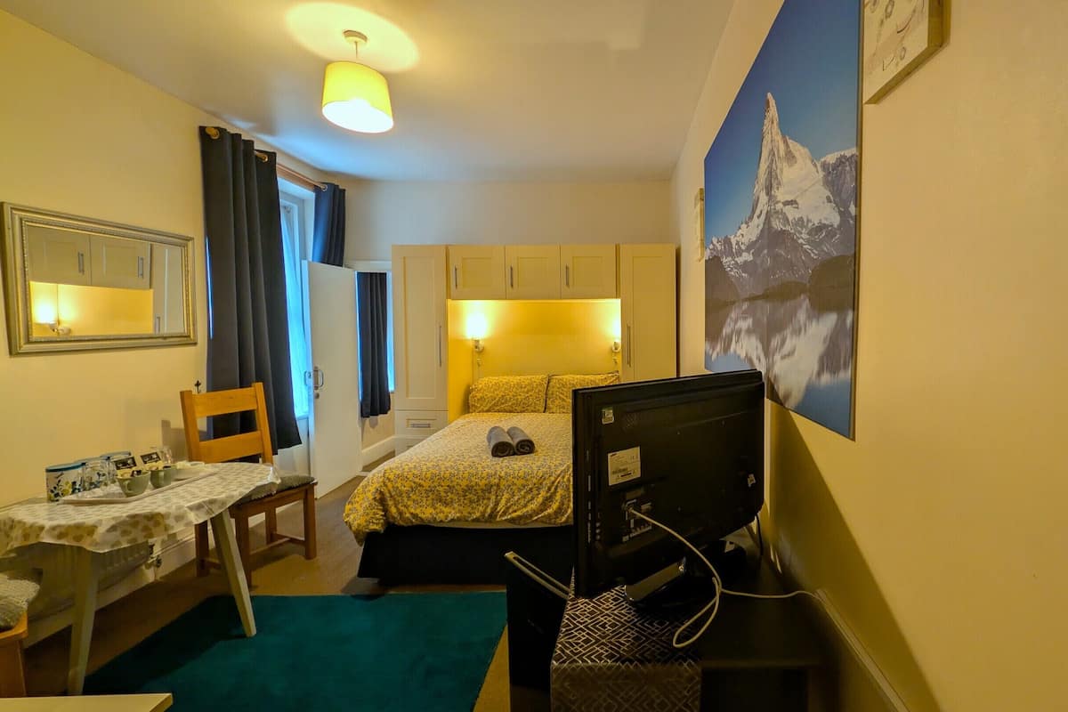 Habitación de hotel pequeña con una cama, un escritorio, una silla, una lámpara de escritorio, una computadora y un cuadro de una montaña.