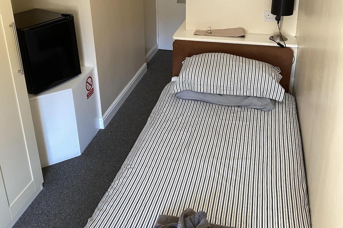 Una habitación de hotel con una cama, un escritorio con lámpara, un refrigerador pequeño y un armario.