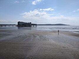 Una playa con un muelle que se adentra en aguas tranquilas, una colina distante y un cielo despejado.