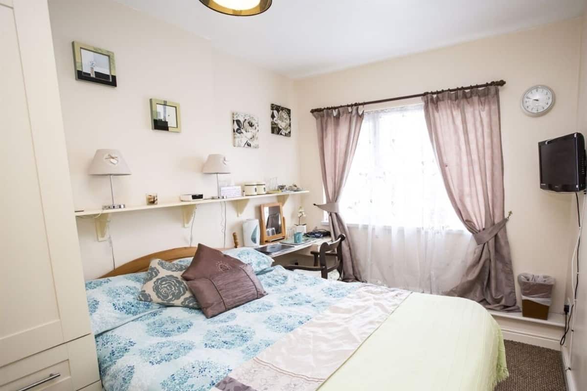 Un dormitorio con cama, mesitas de noche, escritorio, silla, reloj y ventana con cortinas.
