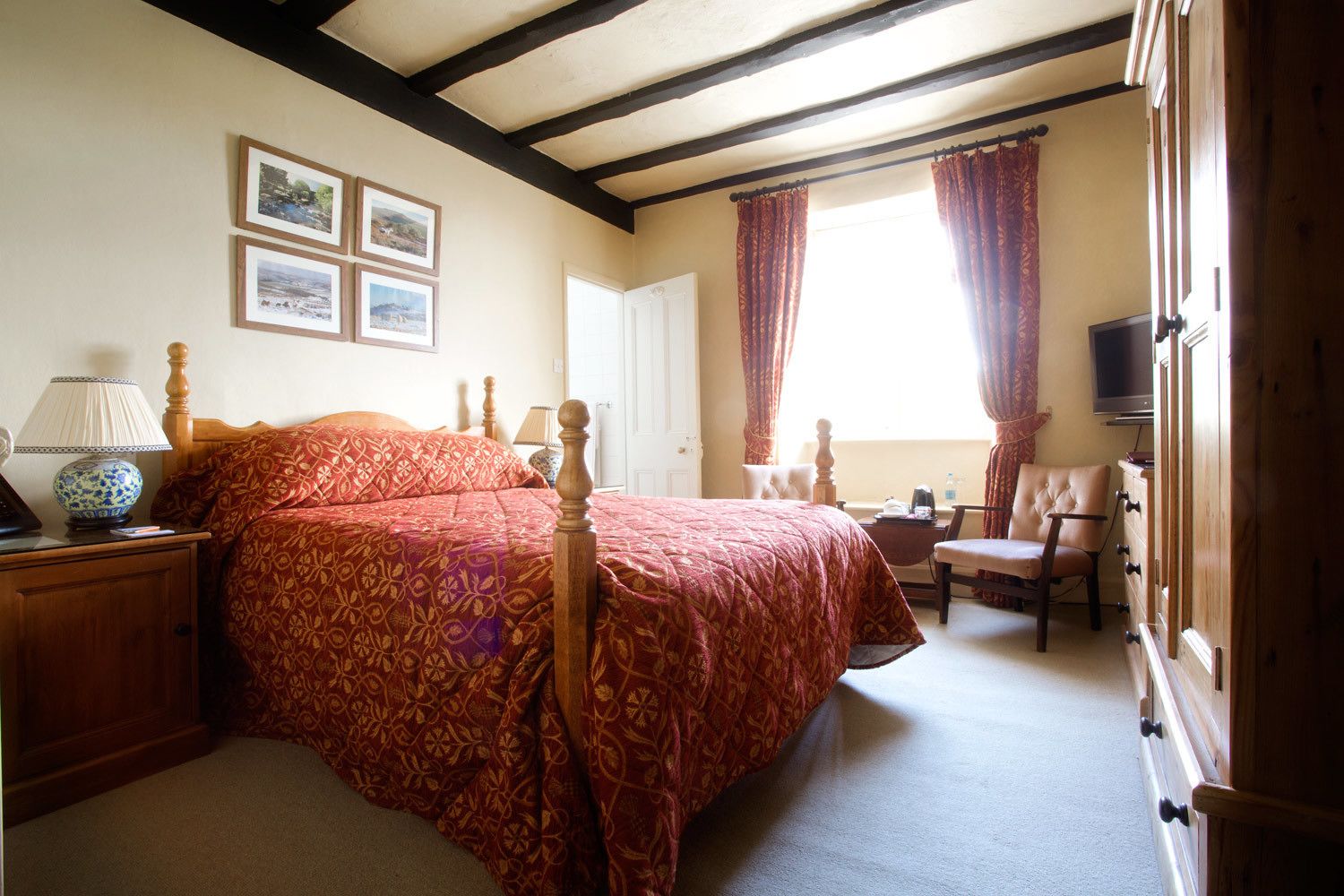 A bedroom with a large bed, a wooden nightstand, a lamp, a chair, a dresser, a television, and a window with curtains.