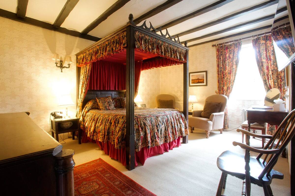 A four-poster canopy bed with a floral bedspread, a wooden desk with a chair, and a red carpet.