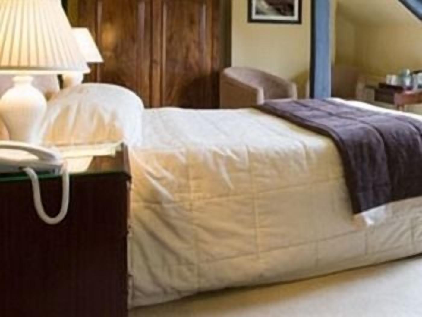 A neatly made bed with a white comforter and a brown throw blanket. There is a bedside table with a lamp and a telephone.