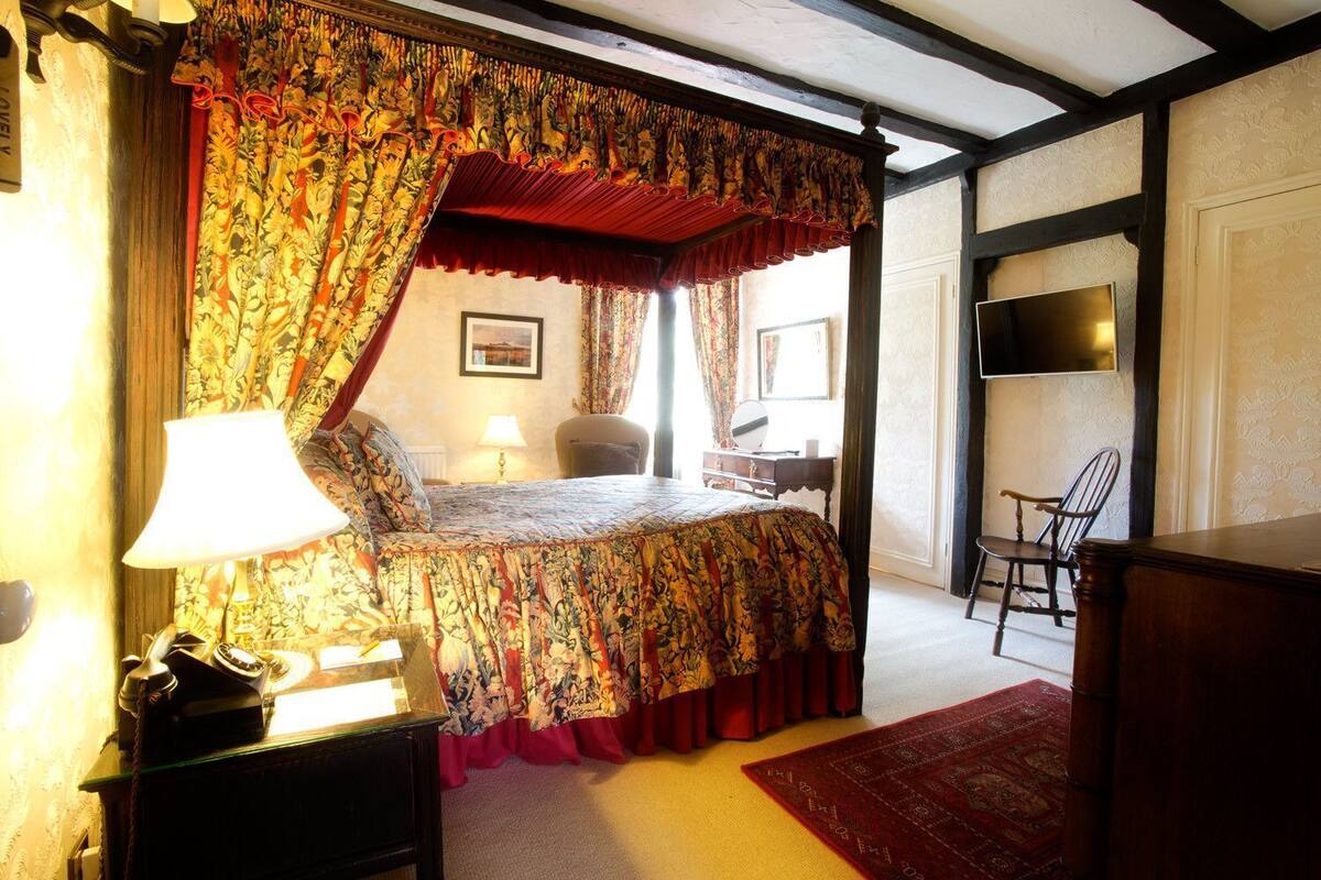A four-poster bed with floral bedspread, a desk with a chair, and a mirror on the wall.