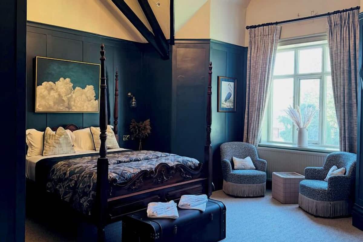 A bedroom with a four-poster bed, a large window with curtains, a chair, and a small table.