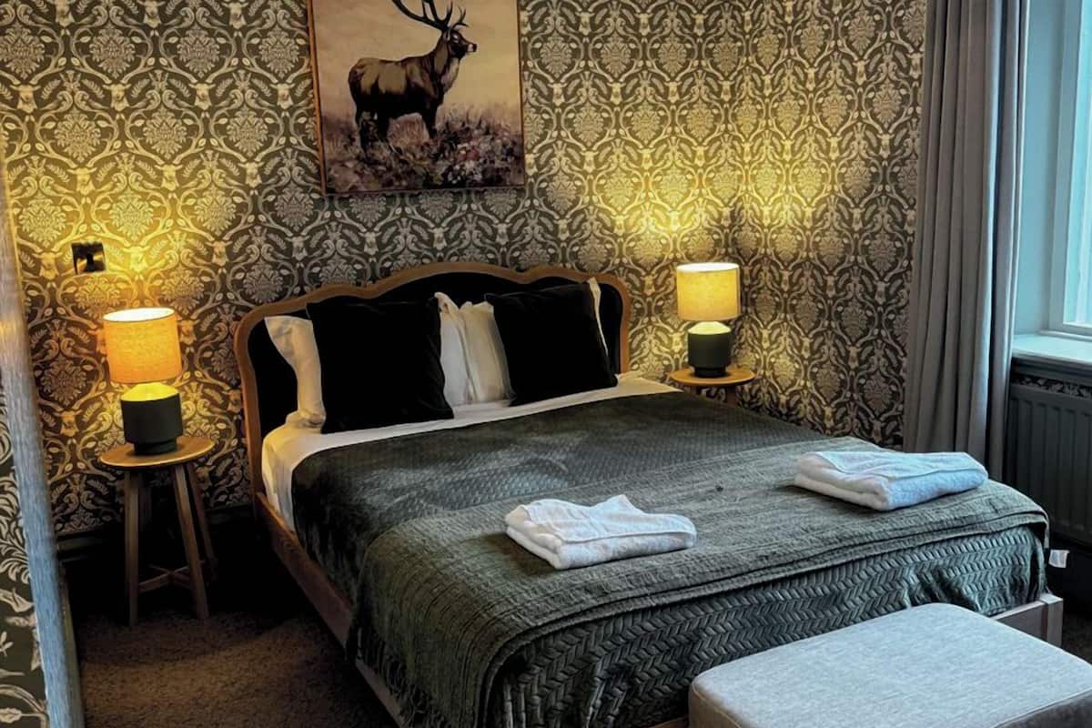 A neatly made bed with a green blanket, two bedside lamps, a painting of an elk, and a patterned wallpaper.