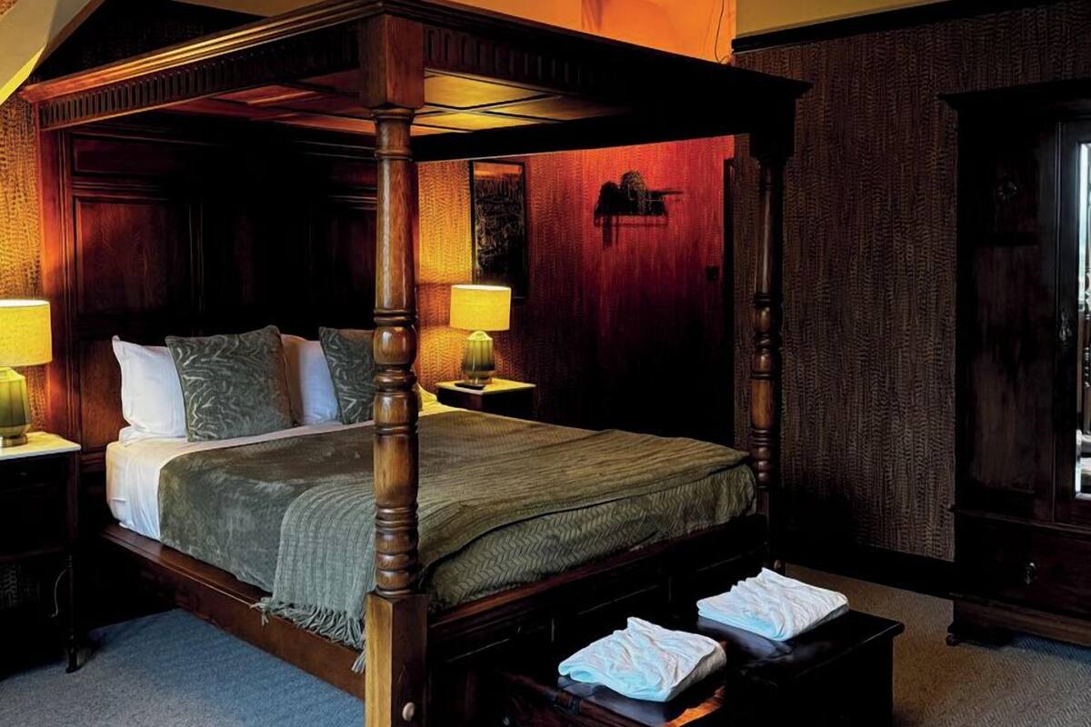 A four-poster bed with a wooden frame, a green bedspread, and two pillows. There are two bedside tables with lamps, and a large mirror on the wall.