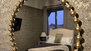 Quarto com um espelho grande e detalhado, cama com cabeceira escura, mesa de cabeceira com abajur e janela com vista para edifícios.