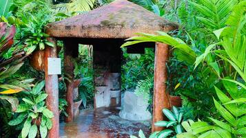 Escape to a serene tropical oasis beneath a thatched roof, surrounded by lush vegetation and a gentle stream.