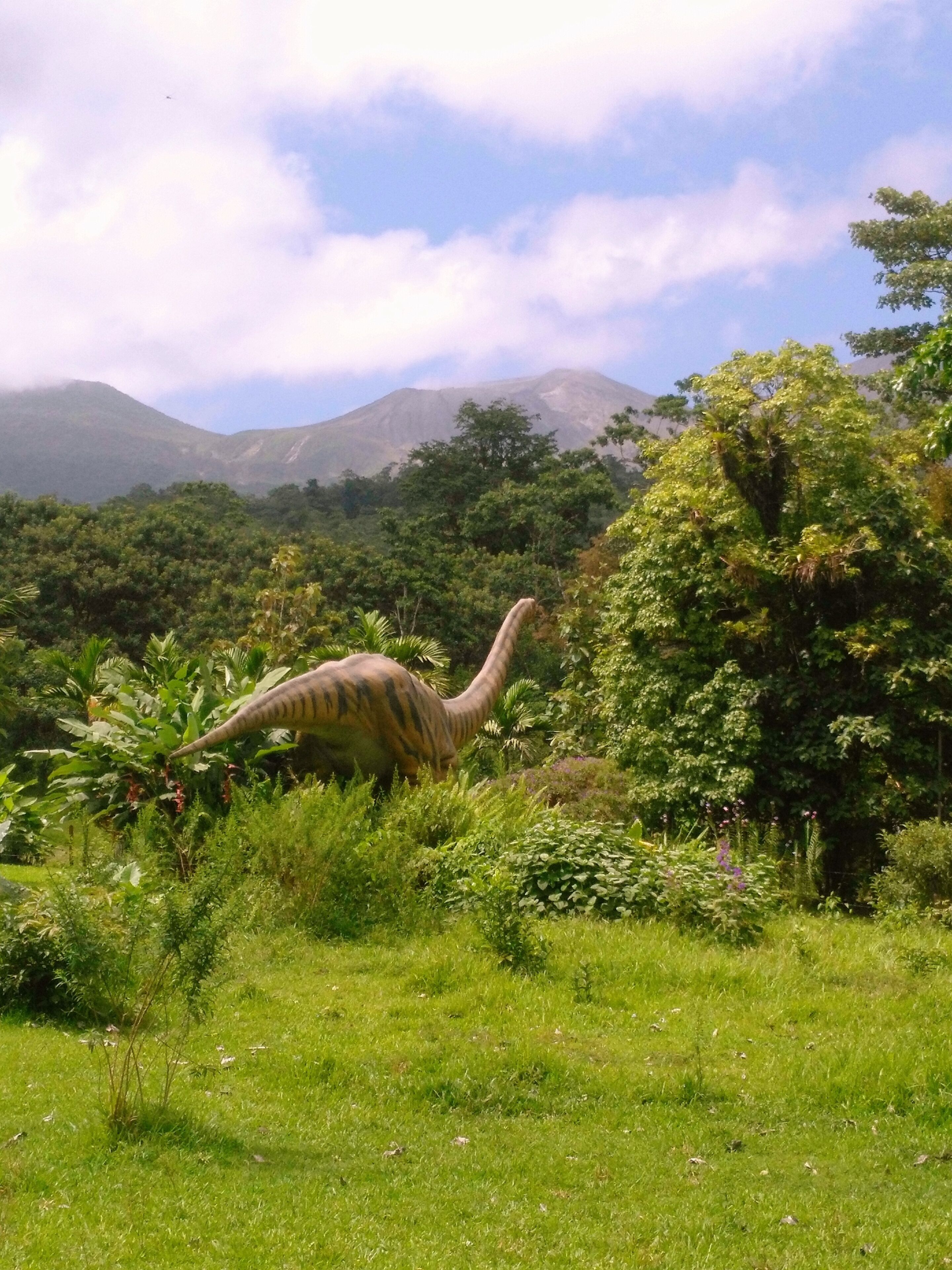 Un modelo de dinosaurio en un paisaje verde exuberante con montañas al fondo.