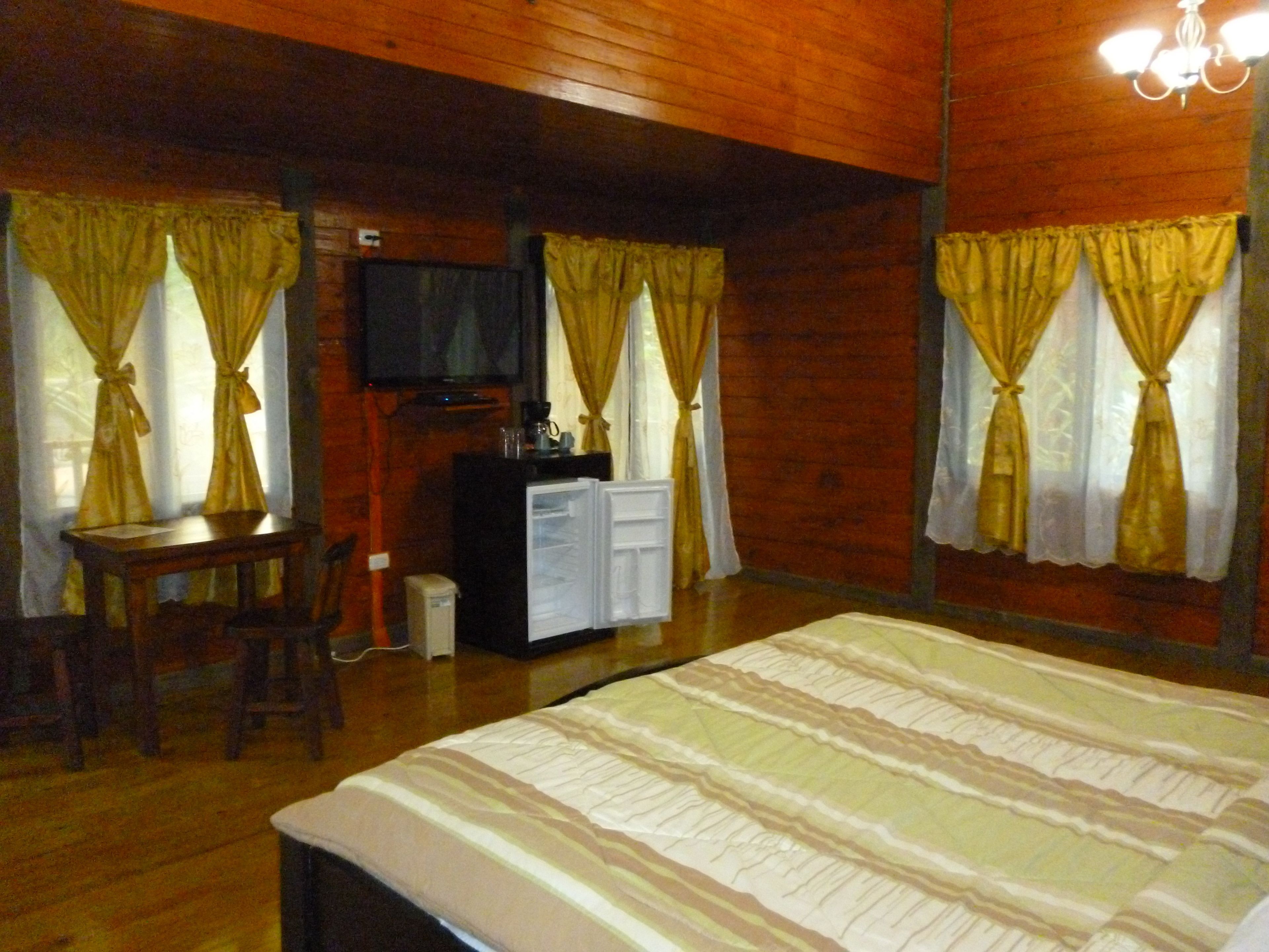 A room with wooden walls, a bed with a striped bedspread, a desk with a chair, a television, and a small cabinet.