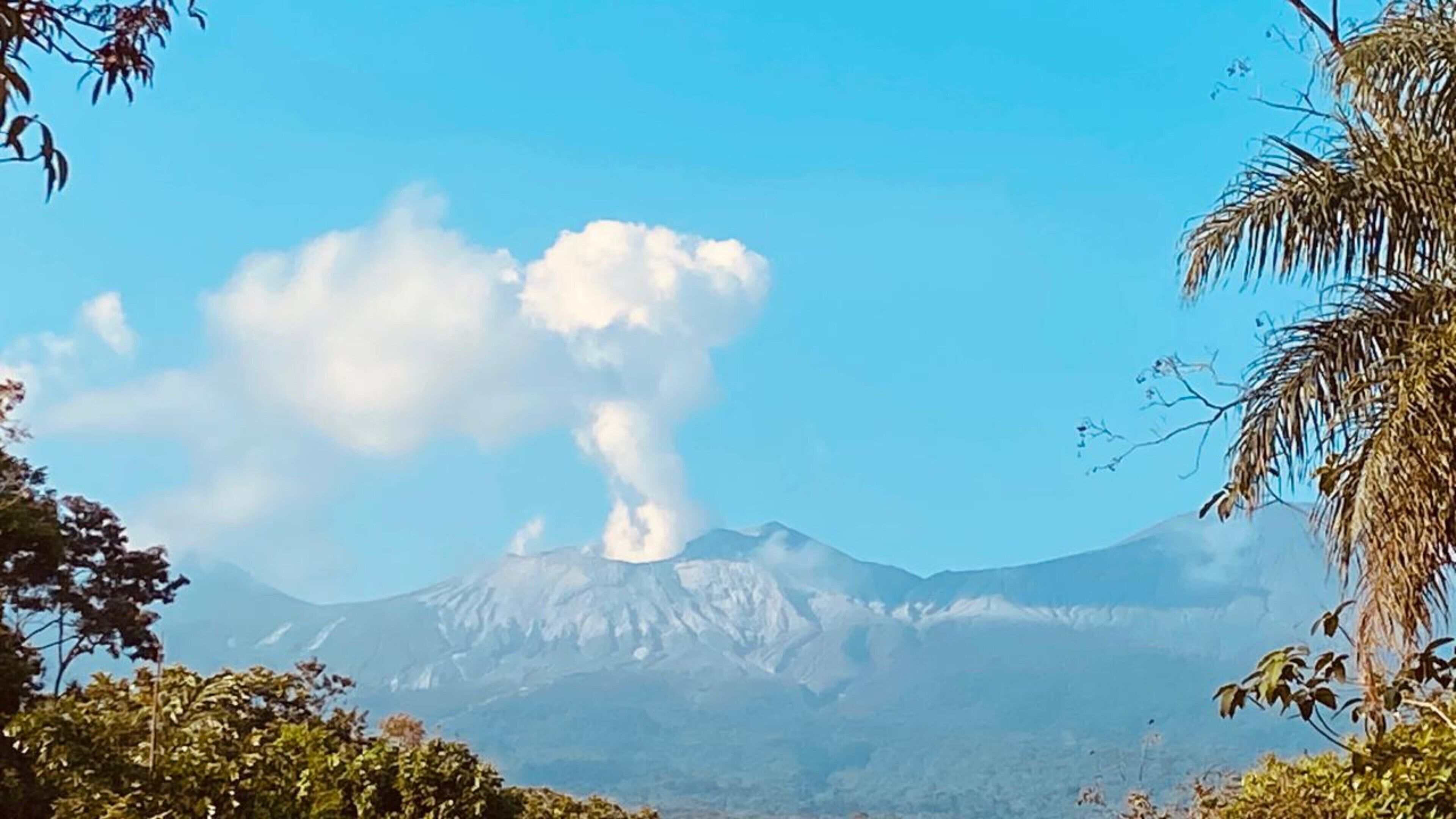 Una montaña con una columna de humo visible, rodeada de árboles y cielo despejado.