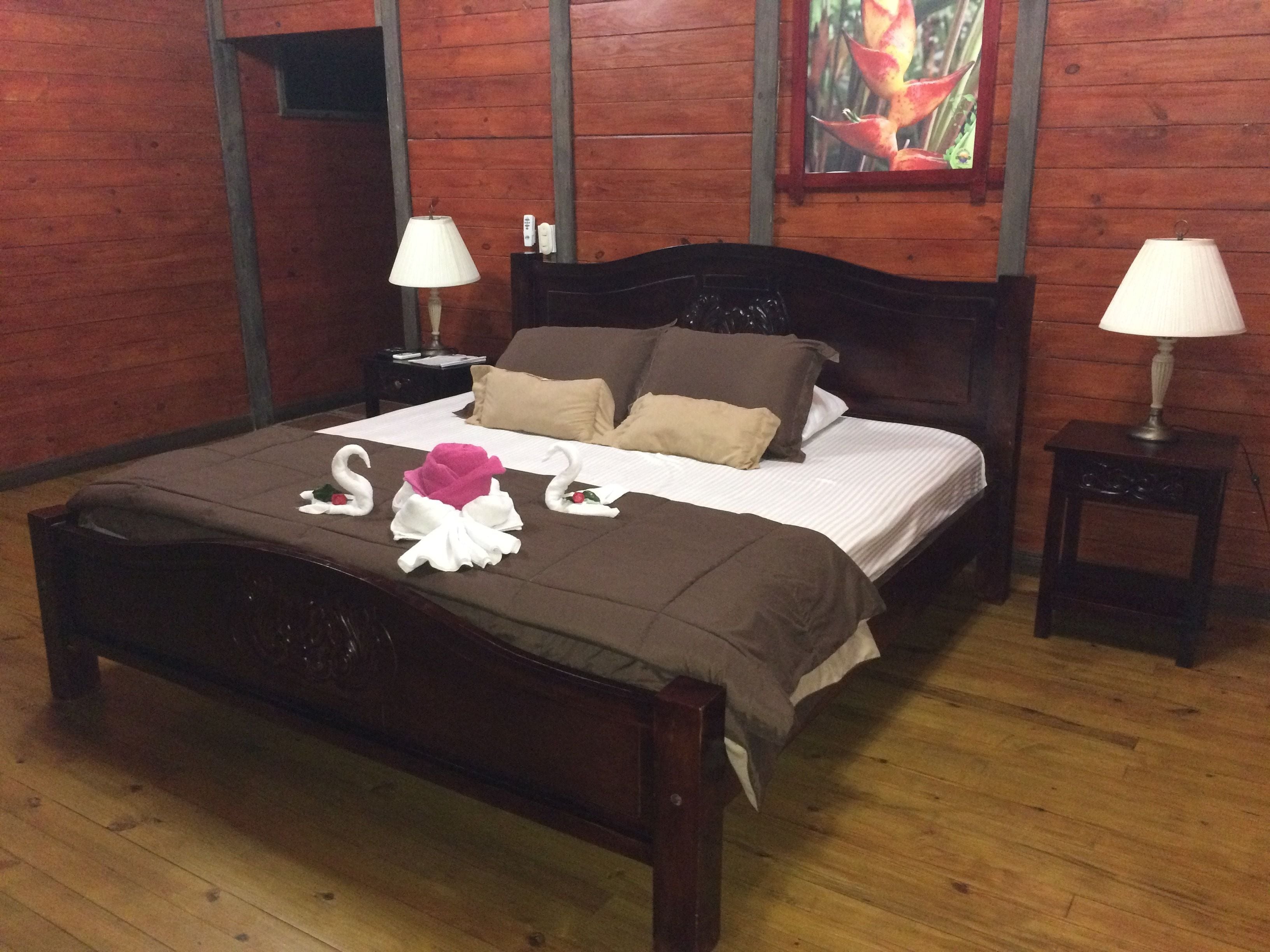 Una cama bien hecha con una colcha marrón y almohadas, dos mesitas de noche con lámparas, y un cuadro en la pared.