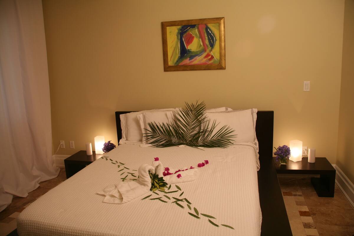 Un lit bien fait, recouvert d’une courtepointe blanche, agrémenté d’une serviette décorative et d’un bouquet de fleurs. Deux tables de chevet, ornées de bougies, se trouvent de part et d’autre du lit, et un tableau est accroché au mur, au-dessus de la tête de lit.