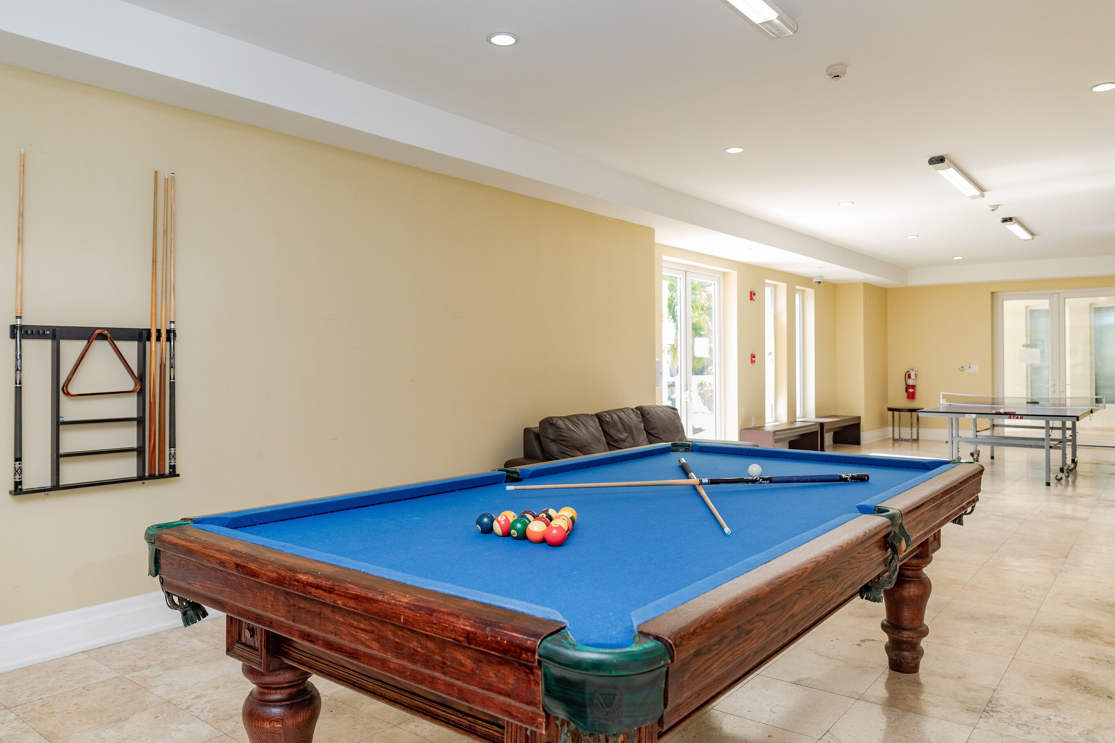 A pool table with balls, a sofa, and a billiards rack in a room with beige walls and a tiled floor.