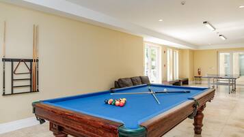 A pool table with balls, a sofa, and a billiards rack in a room with beige walls and a tiled floor.