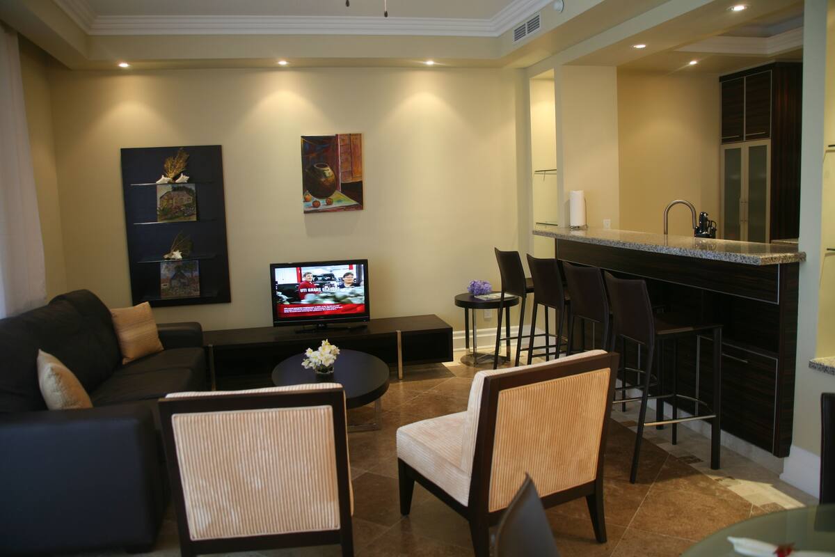 Un salon moderne avec un canapé, des fauteuils, une table basse et un téléviseur. On aperçoit un coin cuisine à l’arrière-plan.