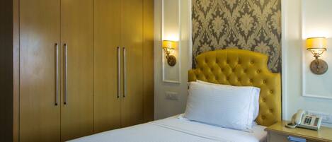 Una habitación de hotel con cama, cabecera amarilla, mesita de noche con teléfono y armario de dos puertas.
