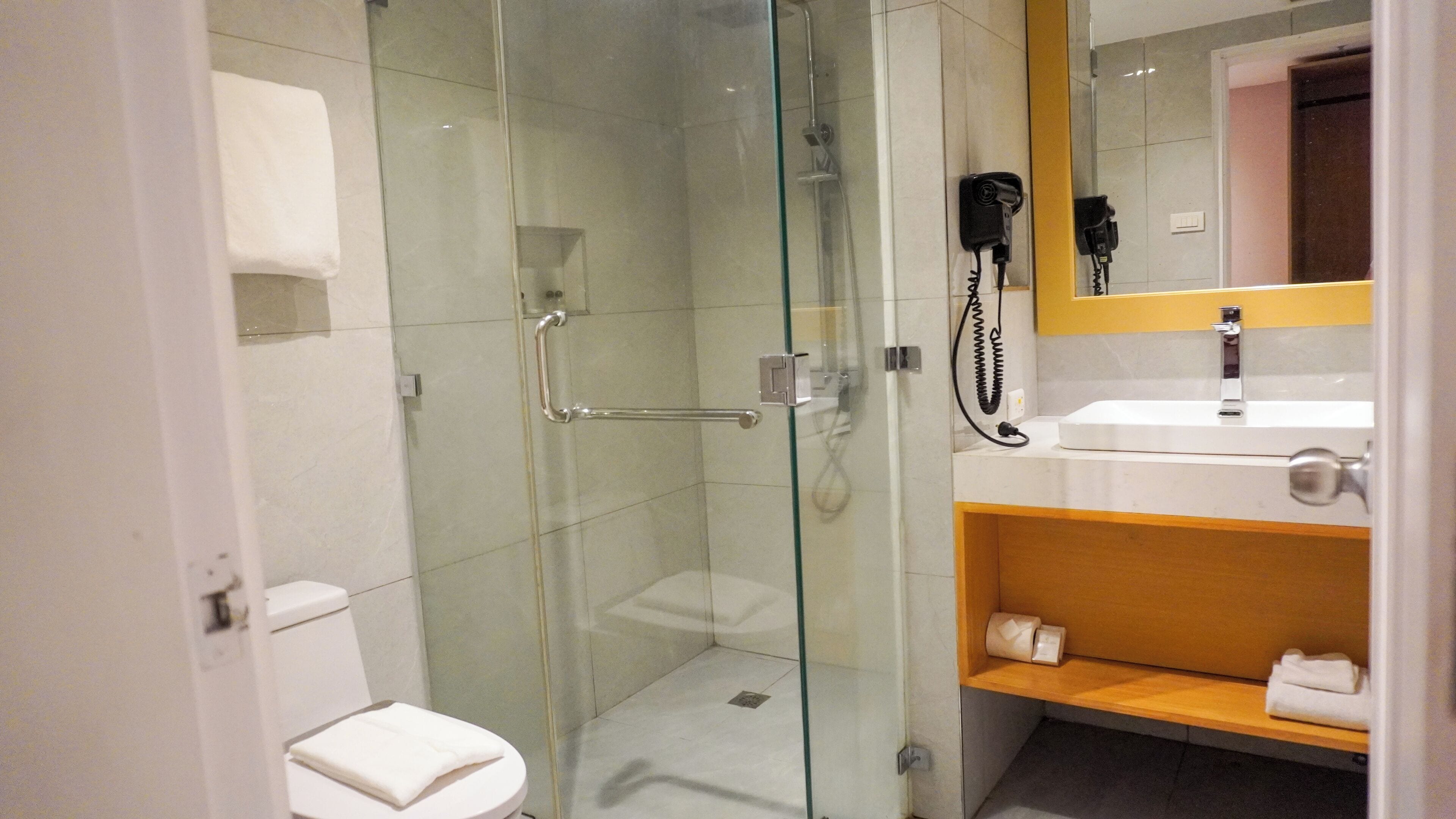 A modern bathroom with a glass-enclosed shower, a white toilet, a sink with a mirror, and a wooden shelf with towels.