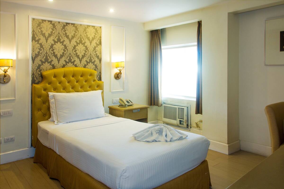 A hotel room with a large bed, a headboard, two bedside lamps, a telephone, a window with curtains, and a wall-mounted air conditioner.