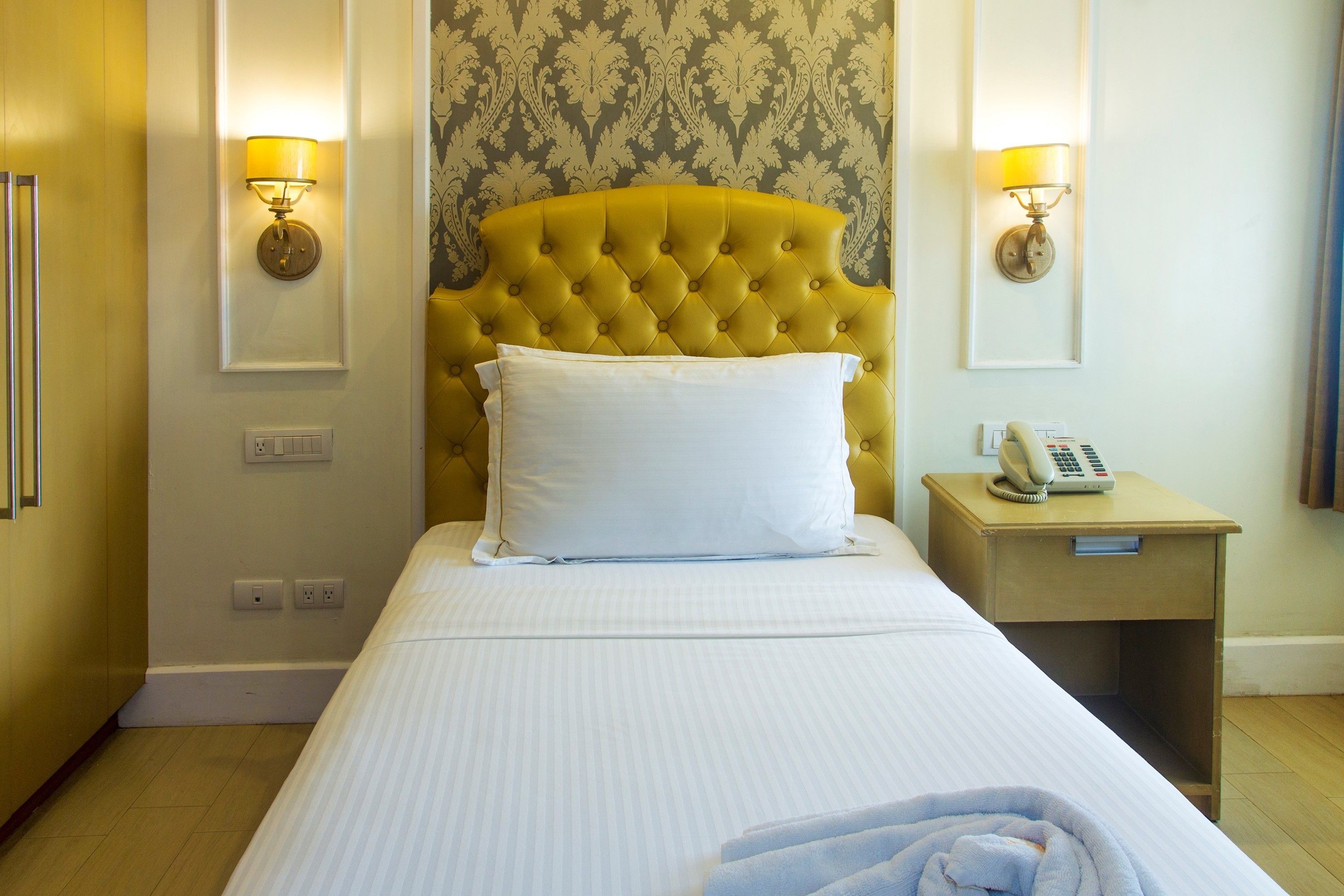 A hotel room with a tufted headboard, a bed with white linens, two wall-mounted lamps, a bedside telephone, and a small wooden nightstand.