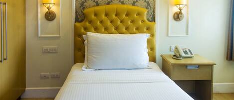 A hotel room with a tufted headboard, a bed with white linens, two wall-mounted lamps, a bedside telephone, and a small wooden nightstand.