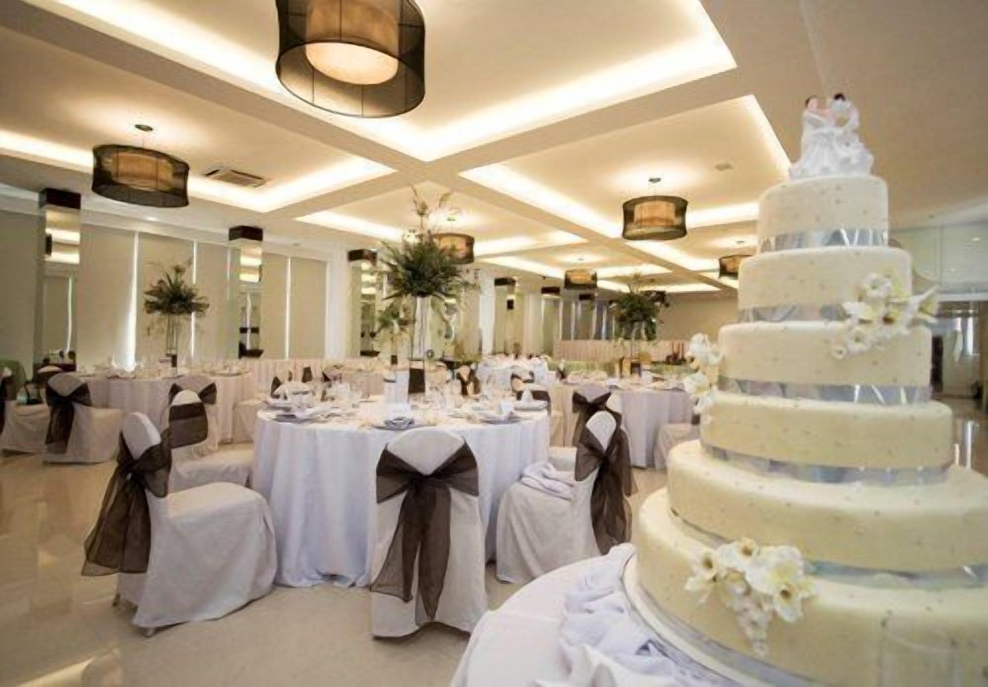 A wedding reception hall with round tables, white tablecloths, and chairs with brown sashes. A multi-tiered wedding cake is prominently displayed in the foreground.