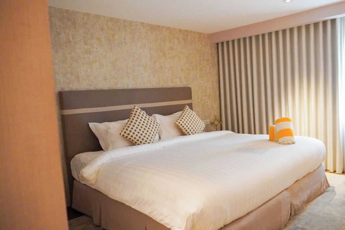 A large bed with a headboard, multiple pillows, and a rolled towel on the foot of the bed.