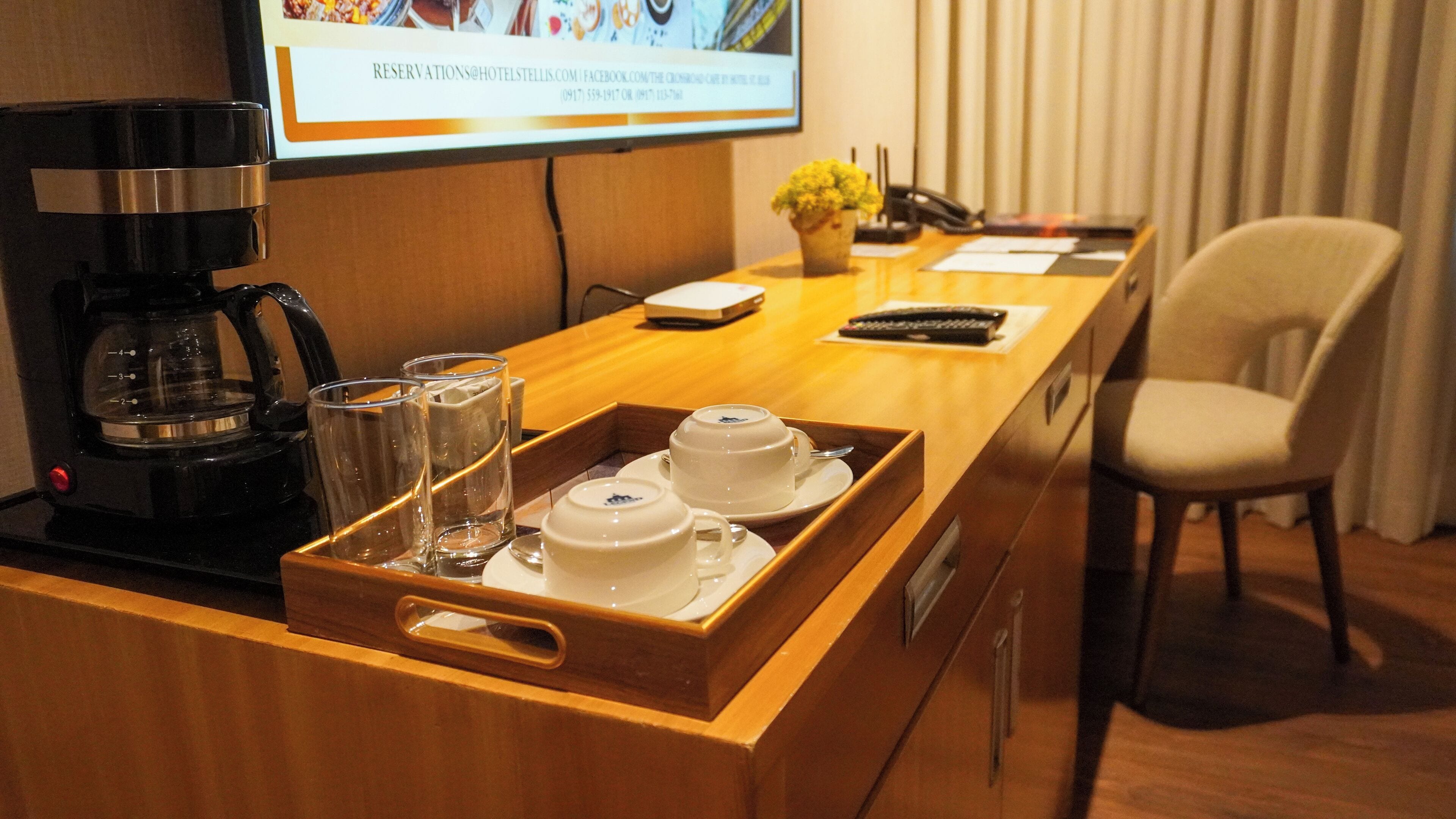 Habitación de hotel con un escritorio de madera, una cafetera, una bandeja con tazas de té y vasos, un teléfono y una silla.