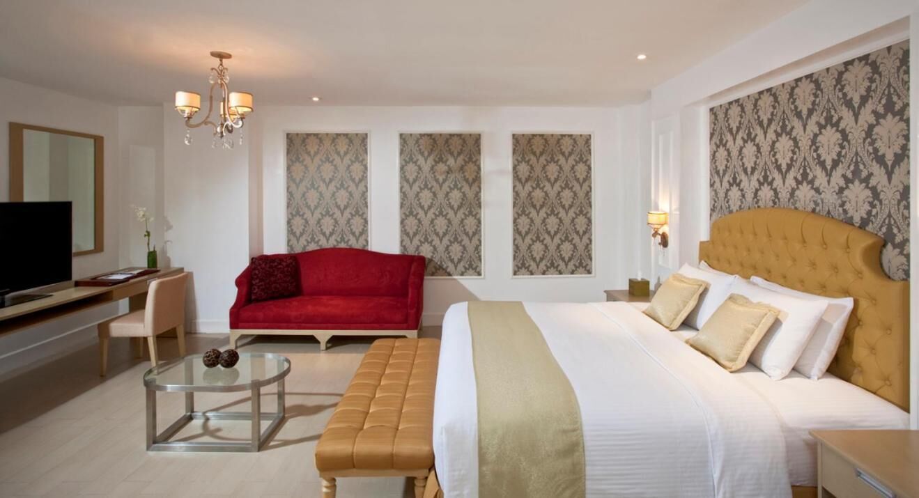 A hotel room with a large bed, a red sofa, a glass coffee table, a television, and patterned wallpaper.