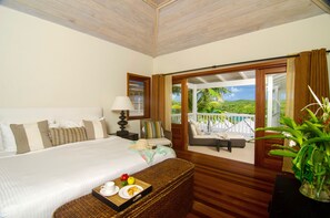 A hotel room with a large bed, a small table with breakfast items, a wicker bench, and a view of a balcony with palm trees. - Residences at Nonsuch Bay Antigua (Freetown)
