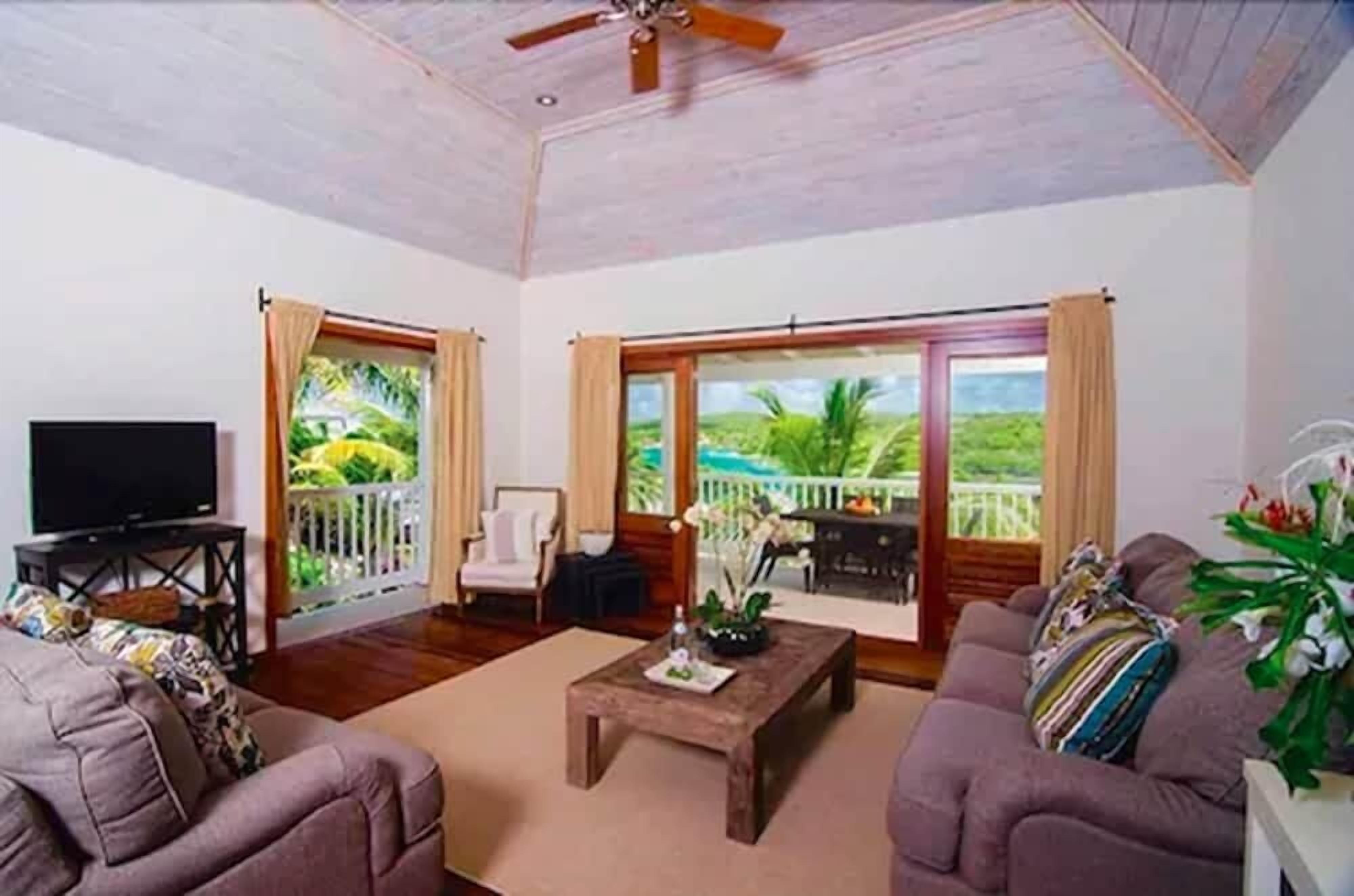 Amplia sala de estar con televisor, dos sofás, una mesa de centro y ventanas grandes que dan a vistas de vegetación y un balcón.