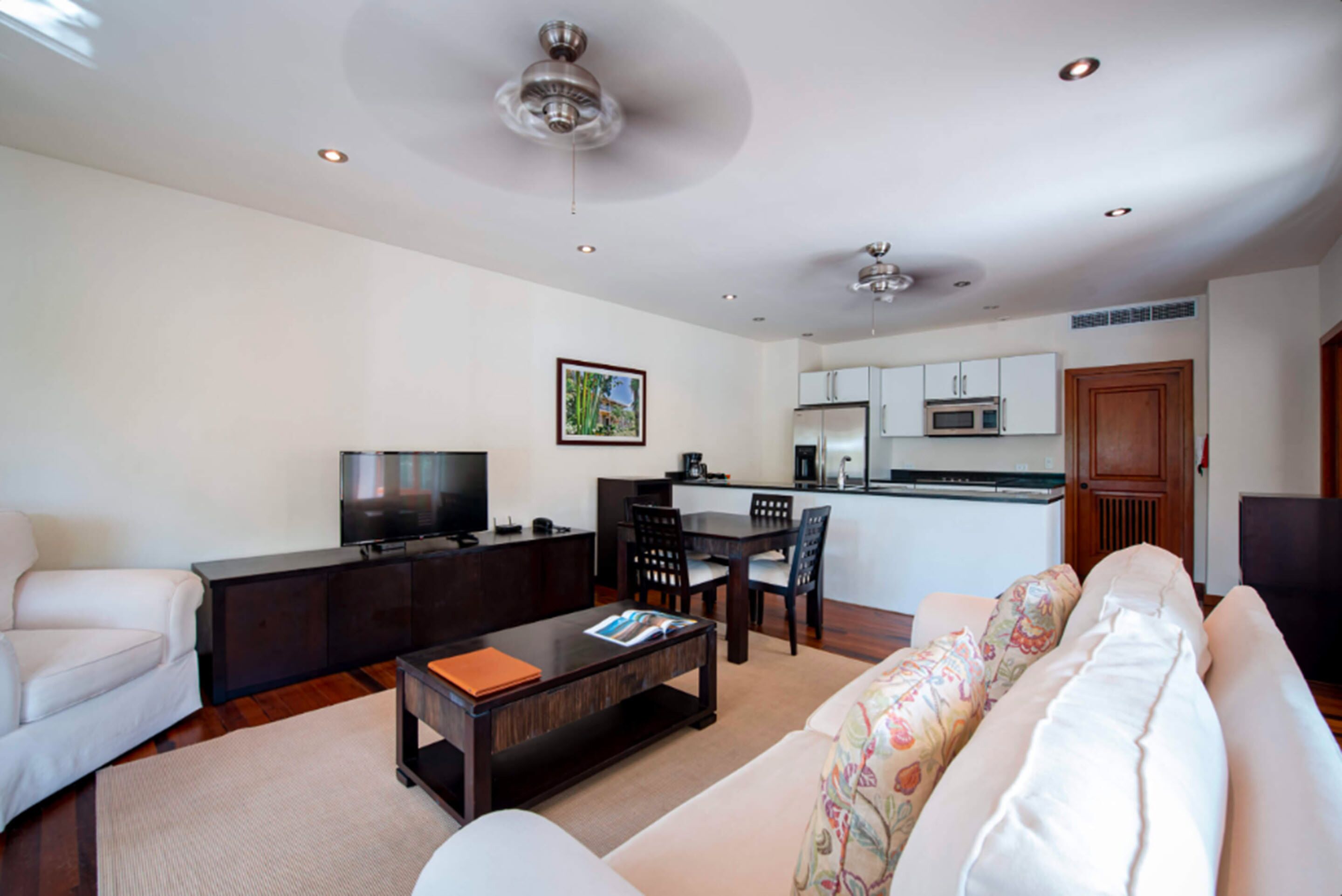A modern living room with a sofa, coffee table, TV, and a dining area with chairs and a kitchen in the background.