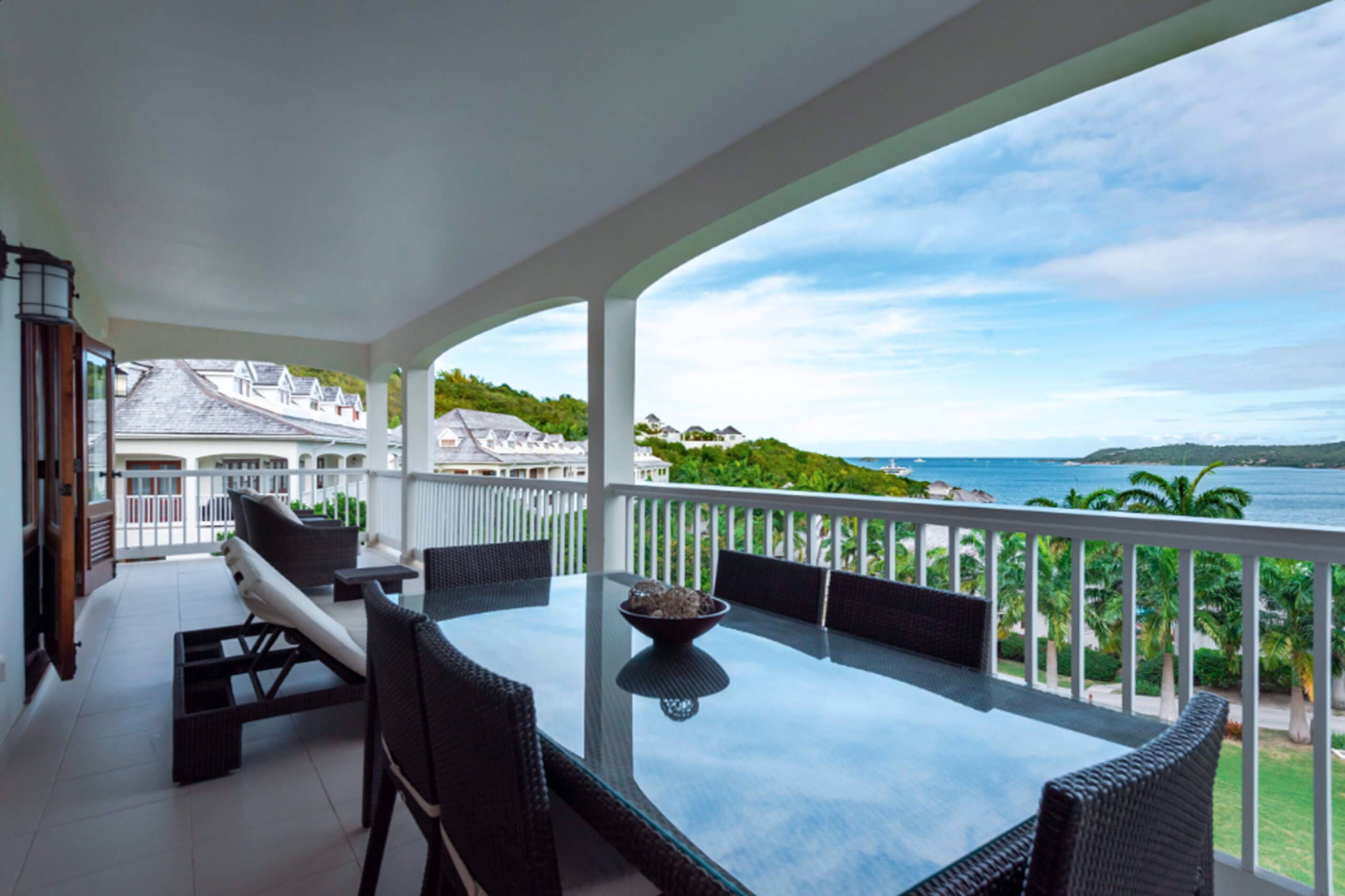 Un balcon doté d’une table en verre et de chaises en osier, offrant une vue sur la côte, avec des maisons et de la verdure.