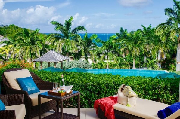 A poolside area with wicker furniture, a table with a bottle and glasses, and a view of the ocean and palm trees. - Residences at Nonsuch Bay Antigua (Freetown)