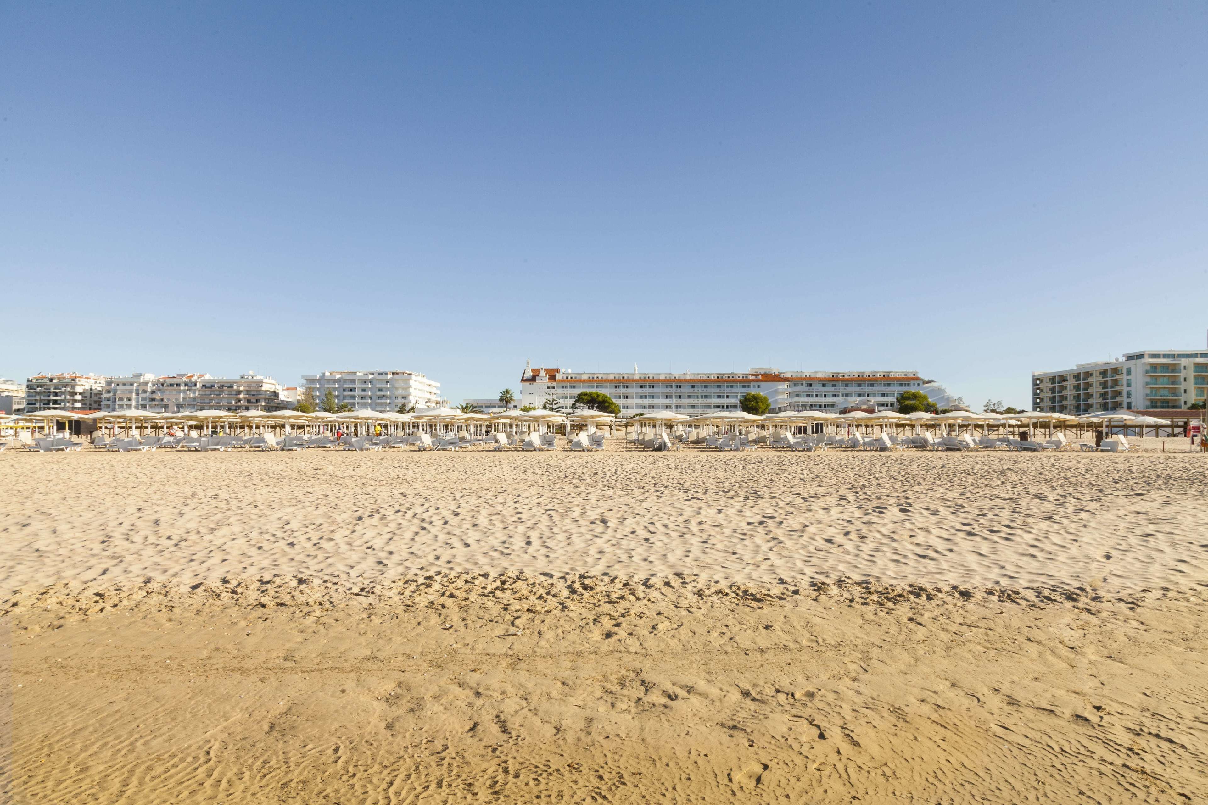 A beach with rows of sun loungers and umbrellas, a large building with multiple floors, and several smaller buildings in the background.