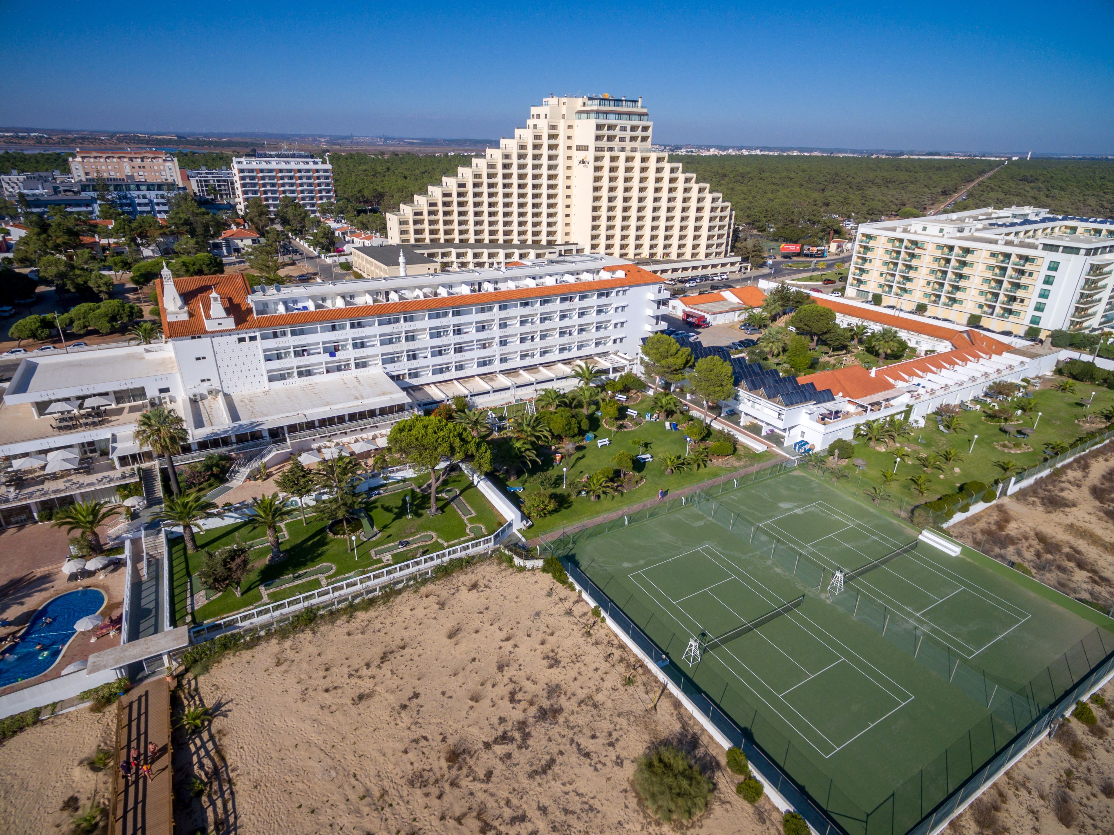 Vista aérea de un complejo hotelero con varias edificaciones, una cancha de tenis y zonas verdes circundantes.